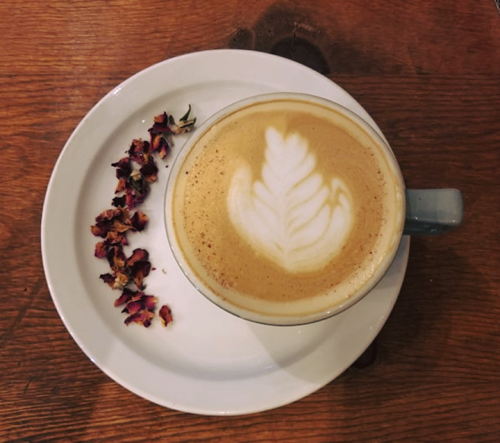 A latte with a leaf design in foam, served on a plate with dried rose petals beside it, on a wooden table.