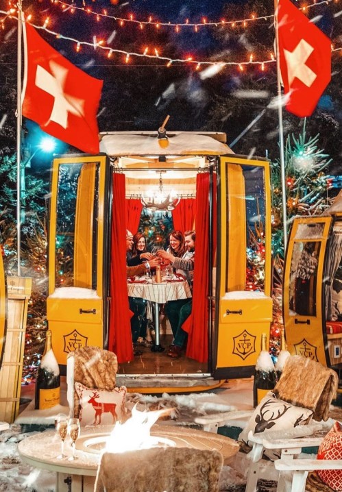 A cozy outdoor setting with a yellow cabin, festive lights, and people enjoying drinks by a fire, surrounded by snow.