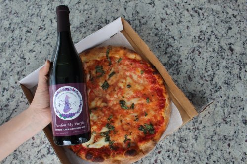 A hand holding a bottle of wine next to a pizza in a takeout box on a speckled countertop.