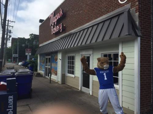 A mascot in a blue jersey poses outside a bakery, with trash bins nearby and a storefront in the background.