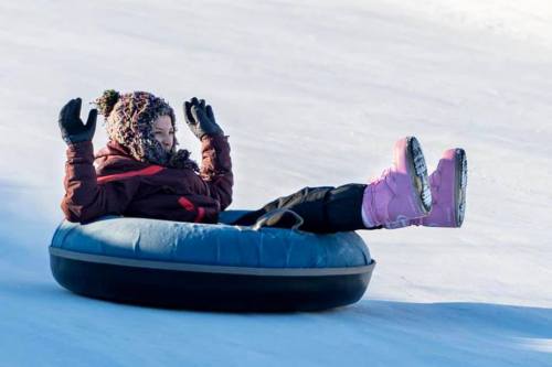 A child in winter gear joyfully rides a snow tube down a snowy slope, hands raised in excitement.