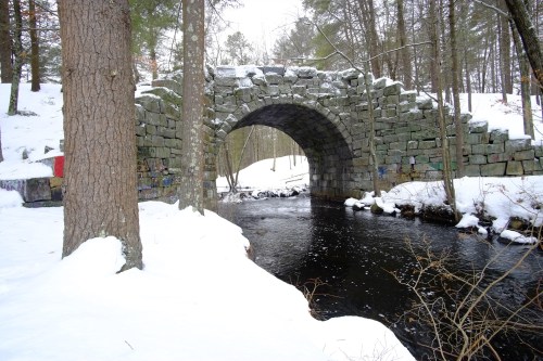 A stone arch bridge over a snowy stream, surrounded by trees in a winter landscape.