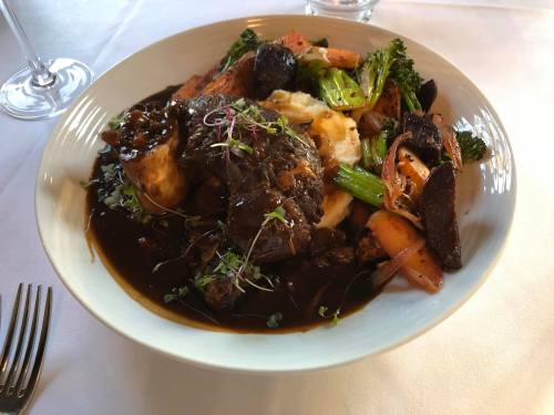 A plate of braised beef with mashed potatoes, roasted vegetables, and a rich sauce, garnished with microgreens.