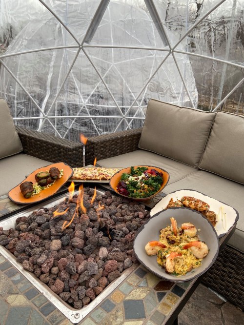 A cozy dining setup with various dishes around a fire pit inside a geodesic dome.