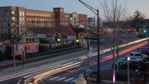 A city street at dusk, featuring light trails from vehicles, historic buildings, and traffic signals.