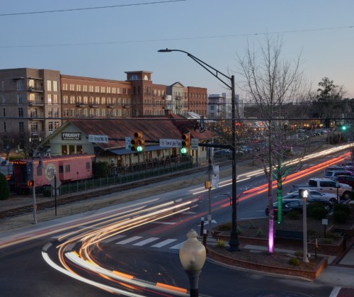 A city street at dusk, featuring light trails from cars, historic buildings, and traffic signals.