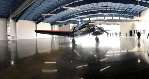A vintage airplane is displayed in a spacious hangar with polished floors and a blue ceiling. People are visible in the background.