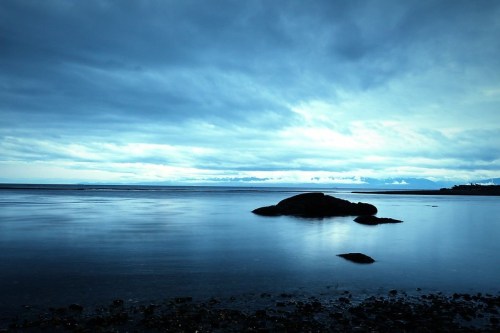 A serene seascape at dusk, featuring calm waters, a large rock, and a cloudy blue sky.