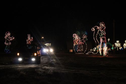 A car drives through a festive display of colorful holiday lights at night, featuring animated figures and decorations.