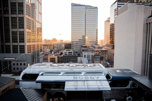 A vintage bus named "Bobby" parked on a rooftop with a city skyline in the background during sunset.