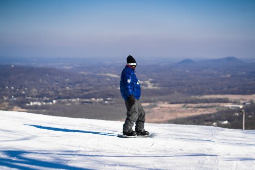 A snowboarder in a blue jacket and black beanie stands on a snowy slope, overlooking a mountainous landscape.