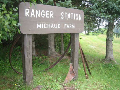 Sign for "Ranger Station" at Michaud Farm, surrounded by trees and grass, with an old tool hanging below.