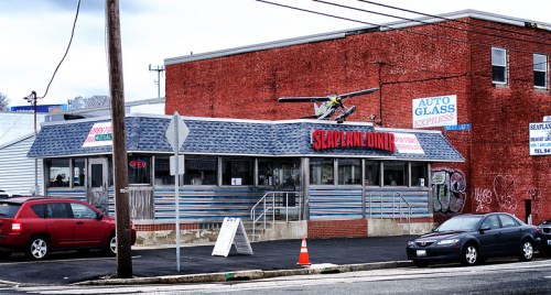 A retro diner with a blue and silver exterior, featuring a small airplane on the roof, surrounded by parked cars.