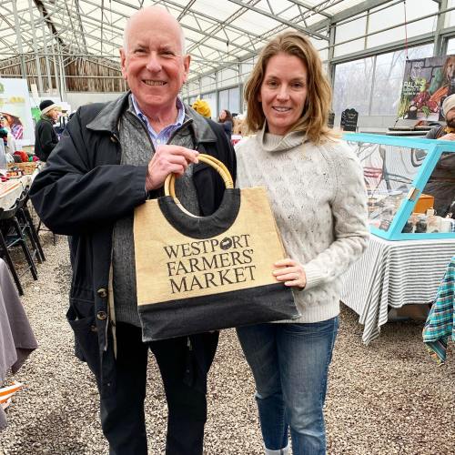 A man and woman smile while holding a Westport Farmers' Market tote bag inside a greenhouse market.