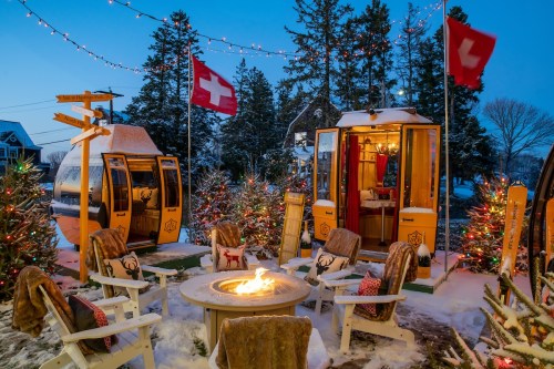 Cozy winter scene with Swiss flags, festive lights, and a fire pit surrounded by snow and decorated trees.