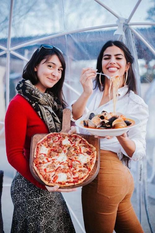 Two women pose with a pizza and a plate of seafood pasta, smiling in a cozy outdoor setting.