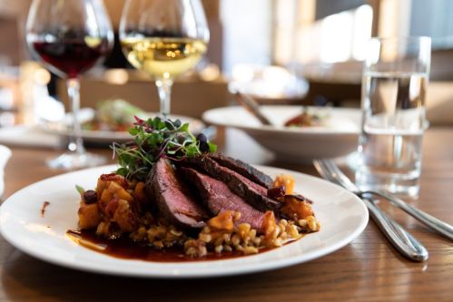 A plate of sliced beef served with vegetables and grains, accompanied by glasses of red and white wine.