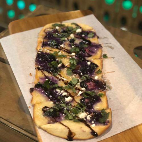 A flatbread topped with purple sauce, crumbled cheese, and herbs, served on a wooden board.