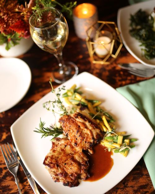 A plate of grilled chicken with sauce, garnished with herbs, served alongside sautéed zucchini and a glass of white wine.