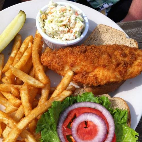 A plate with crispy fried fish on bread, coleslaw, fries, a pickle, and fresh lettuce and tomato slices.