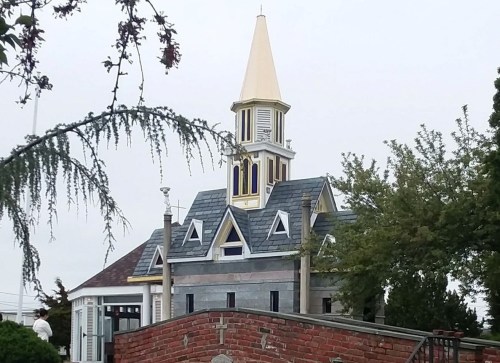 A decorative building with a tall, golden spire and gray shingles, surrounded by trees and a brick wall.