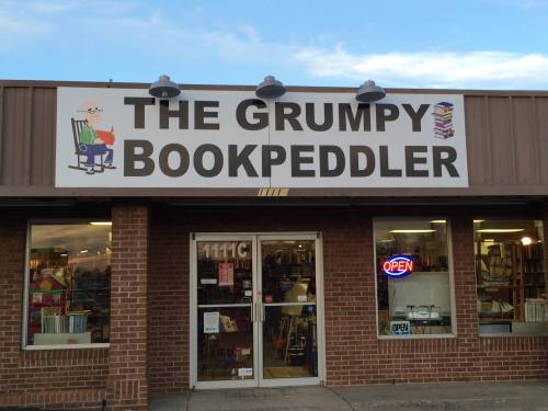 Sign for "The Grumpy Bookpeddler" bookstore, featuring a rocking chair and stacks of books, with an open sign in the window.