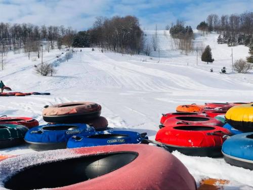 Colorful snow tubes are stacked on a snowy hill, with trees and a slope in the background under a blue sky.