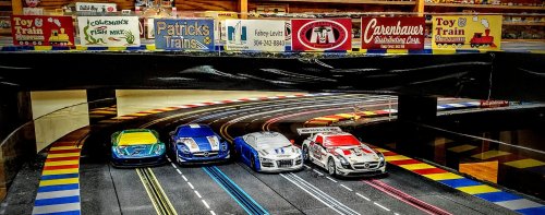 Four colorful toy race cars on a slot car track, with a backdrop of toy store signs and shelves.