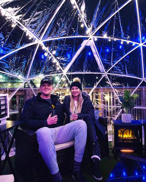 A couple sits inside a cozy, illuminated dome, enjoying drinks by a small electric fireplace.
