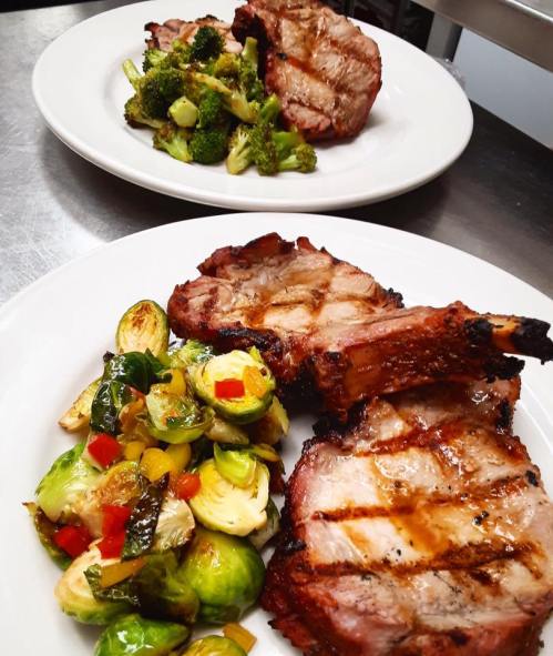 Two plates of grilled pork chops served with sautéed vegetables, including Brussels sprouts and broccoli.