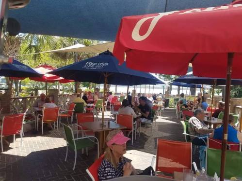 Outdoor dining area with colorful umbrellas, tables filled with people, and a view of greenery and water in the background.
