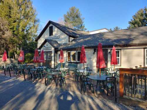 A cozy restaurant exterior with outdoor seating and red umbrellas on a sunny day, surrounded by trees.