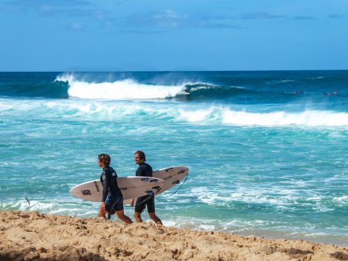 Two surfers walk along the beach carrying their boards, with waves crashing in the background under a clear blue sky.