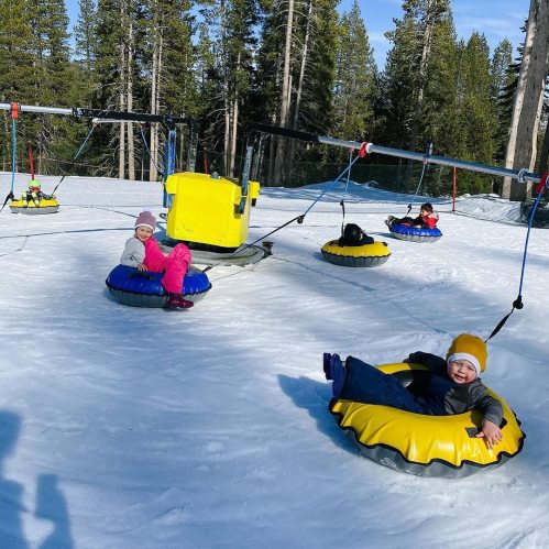 Children enjoying a snow tubing activity on a snowy hill, with trees in the background and a lift in the distance.
