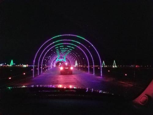 A car drives through a colorful light tunnel with purple and green lights, surrounded by festive decorations at night.