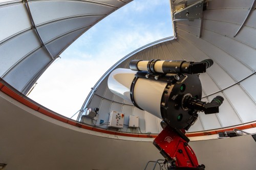 A telescope positioned inside an observatory dome, with an open roof revealing a blue sky.