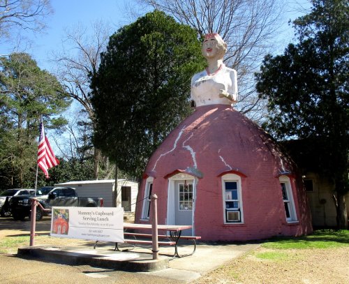 A whimsical building shaped like a woman in a dress, serving as a restaurant, with an American flag nearby.