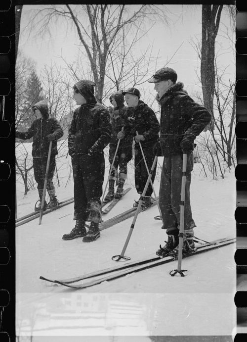 A group of five skiers stands on snow-covered ground, dressed in winter gear, with trees in the background.