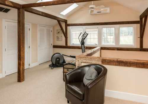 A cozy room with wooden beams, a chair, exercise equipment, and large windows letting in natural light.