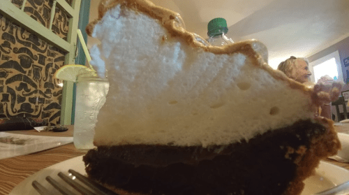 A slice of pie with a chocolate filling and fluffy meringue topping, served on a plate with a drink in the background.
