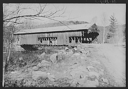 A weathered wooden covered bridge stands on stilts, surrounded by rocks and trees, with mountains in the background.