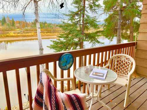 A cozy balcony with a table and chairs, overlooking a calm river and surrounded by trees. A blanket and clock are visible.