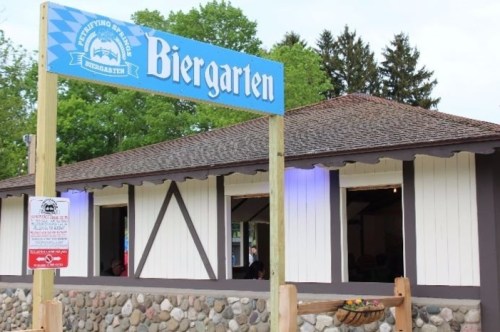 A wooden building with a sign reading "Biergarten," surrounded by greenery and featuring a stone foundation.