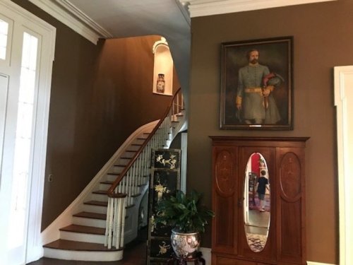 A staircase leads to an upper level, with a large portrait of a man in uniform hanging on the wall beside a wooden cabinet.