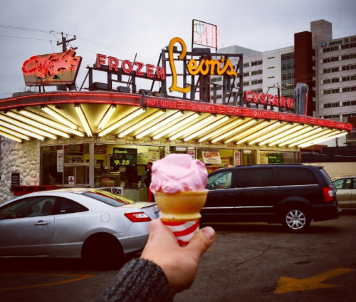 A hand holds a pink ice cream cone in front of Leon's Frozen Custard, featuring a retro neon sign and parked cars.
