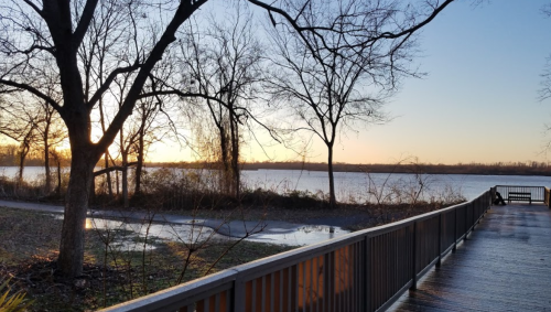 A serene lakeside view at sunset, with bare trees and a wooden walkway reflecting the warm colors of the sky.