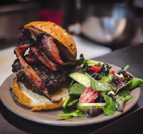 A juicy burger topped with crispy bacon, served on a plate with a fresh salad of greens, cucumbers, and olives.
