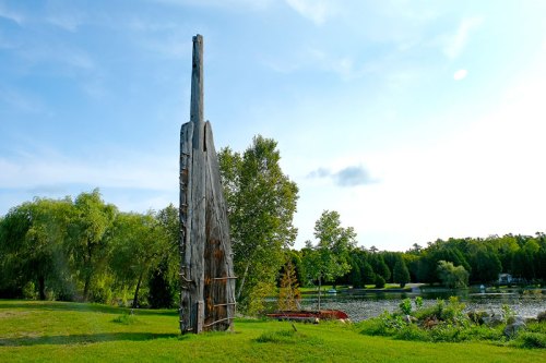 A tall wooden structure stands on a grassy area by a calm lake, surrounded by trees and a clear blue sky.