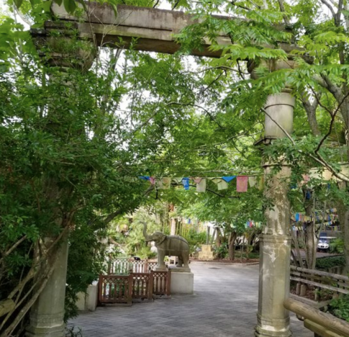 A stone archway surrounded by lush greenery, with a statue of an elephant and colorful prayer flags in the background.
