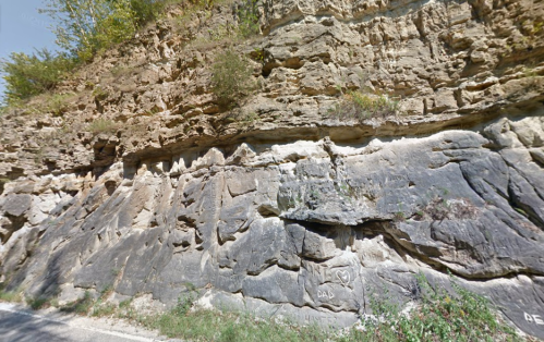 Rocky cliffside with layered stone formations and some vegetation at the top, alongside a narrow road.